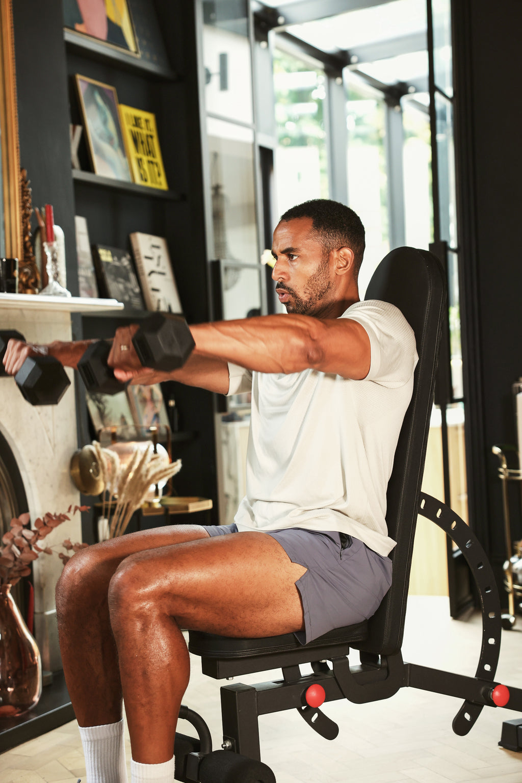 A man in a white t-shirt and gray shorts sits on a black adjustable weight bench, performing a lateral raise with two hex dumbbells in a modern home gym setting.